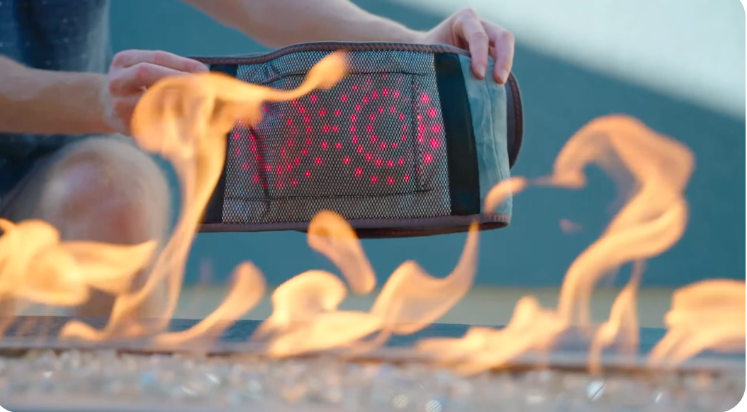 Person haelt GesundHeat Infrarot Heizgurt mit leuchtenden roten LED Lichtpunkten ueber Flammen zur Demonstration von waerme und thermotherapie funktion